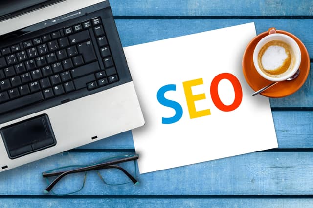 Auf einem alten, blauen Holztisch liegt ein Laptop, eine Brille und ein Blatt mit der Aufschrift SEO