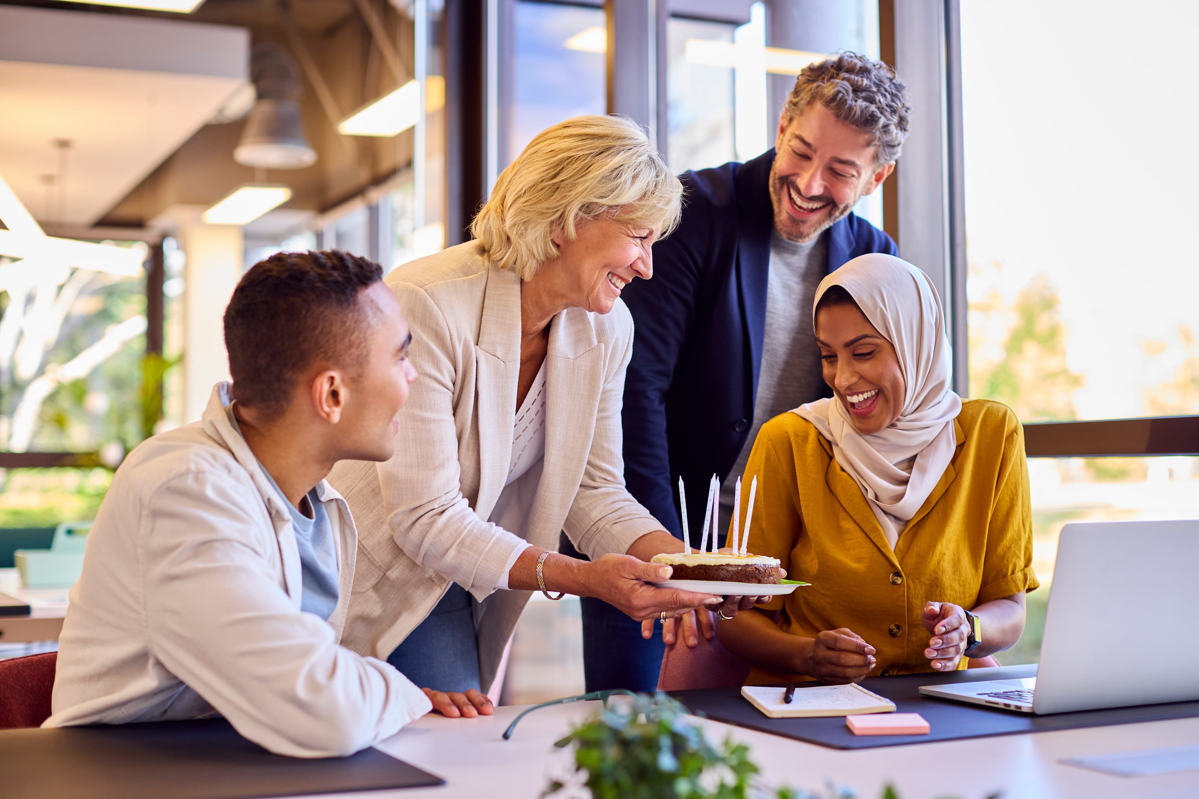 Eine fröhliche, diverse Gruppe von Kollegen feiert gemeinsam einen Geburtstag im Büro – eine Frau überreicht der Kollegin eine kleine Torte mit Kerzen.