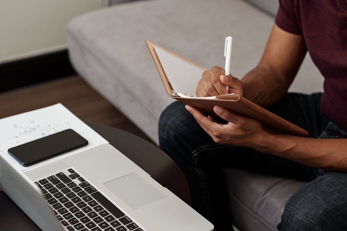 Content Marketing Manager sitzt auf dem Sofa vor seinem Laptop und macht sich Notizen zu Ideen einer neuen Story.