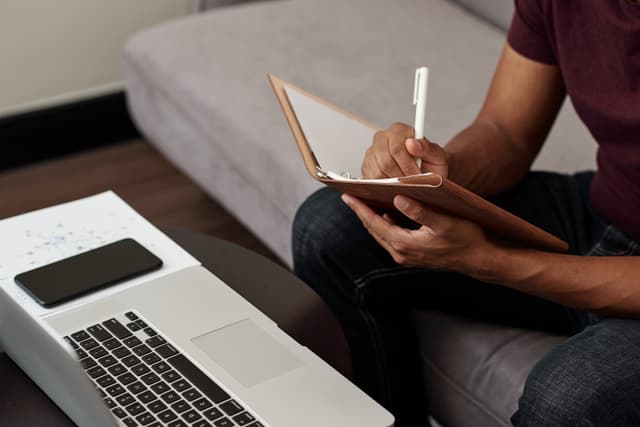 Content Marketing Manager sitzt auf dem Sofa vor seinem Laptop und macht sich Notizen zu Ideen einer neuen Story.