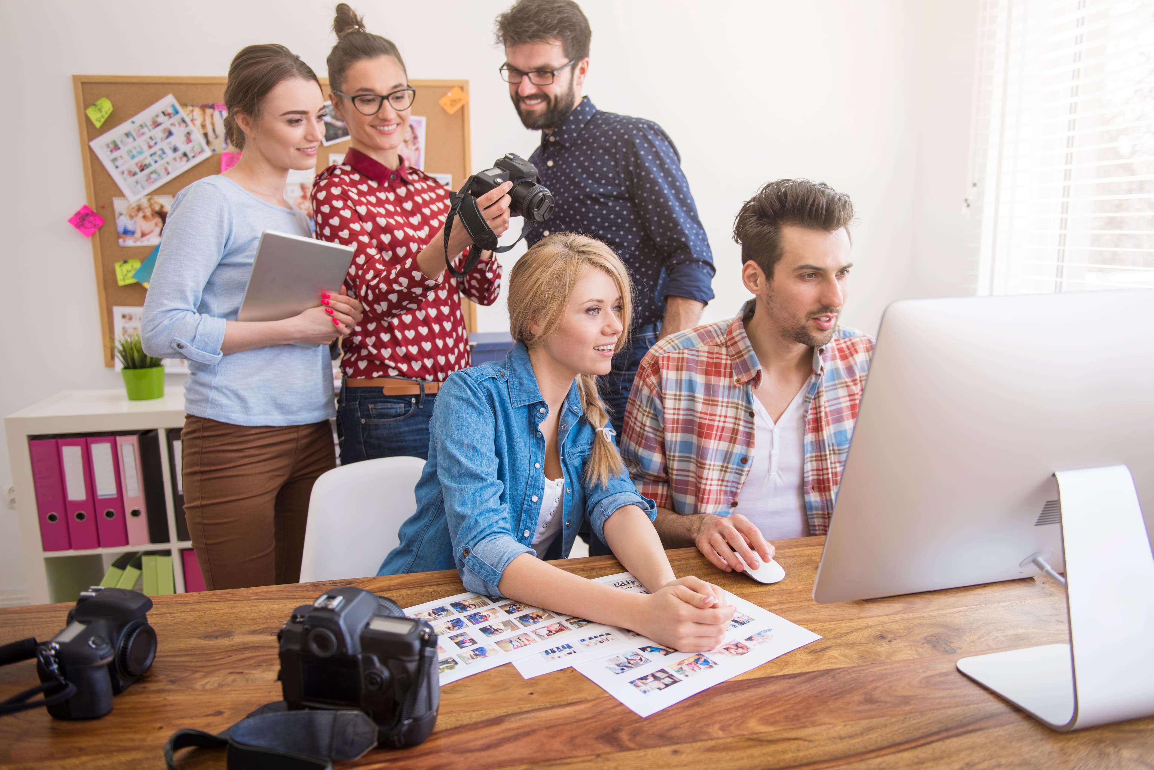 Fünf kreative Teammitglieder besprechen Content-Ideen vor einem großen Monitor mit Kamera-Equipment und Fotoprints.