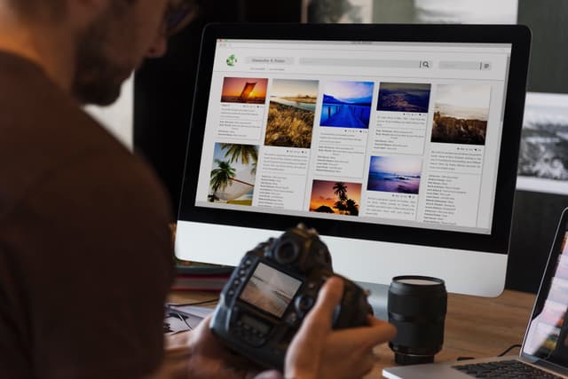 Content Marketing Manager sitzt vor seinem PC mit Kamera in der Hand. Auf dem PC sieht man einen Blog über Fotografie.