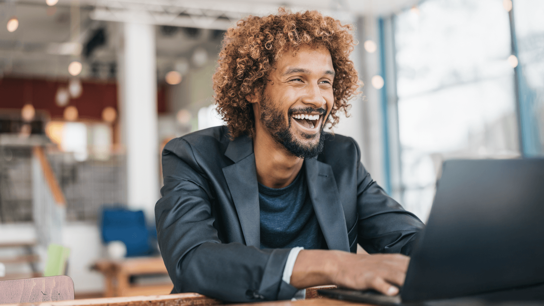 Content Marketing Manager mit Anzug und lockigen Haaren sitzt im Büro am Schreibtisch und lacht.